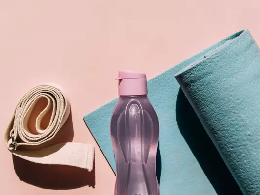 A pink water bottle next to a blue yoga mat and nude yoga strap on a pink background.