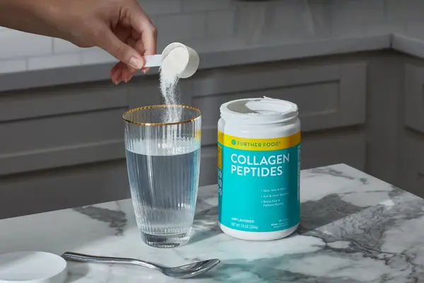 Hand pouring a scoop of Further Food Collagen Peptides Powder into a glass with water next to powder container on a counter