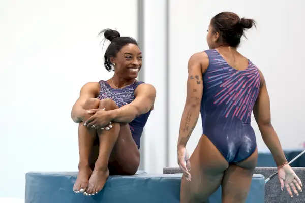Simone Biles le sonríe a Jordan Chiles mientras se sienta en una colchoneta de gimnasia.