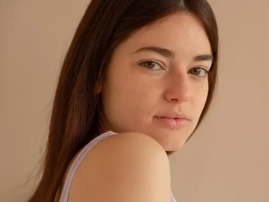 A bare-faced woman with full, fluffy brows looking over her shoulder 