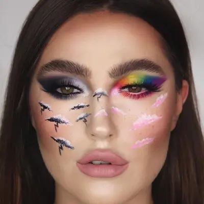Makeup artist wears half-black and white, half-rainbow and pink eye makeup and cheek clouds