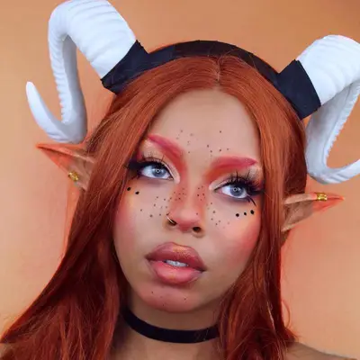 Makeup artist wears an Aries ram-inspired look with red brows, lips, and cheeks and horn headpiece