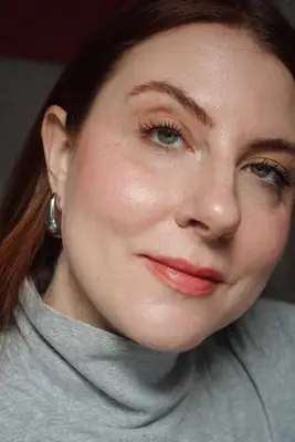 model is posing smiling after just applying a soft peach pink cream blush to the apples of the cheeks