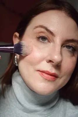 model is applying powder blush with a fluffy brush to apples of the cheeks