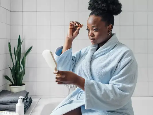 A woman in a bathrobe sitting on the side of the bathtub and tweezing her eyebrows while holding a hand mirror