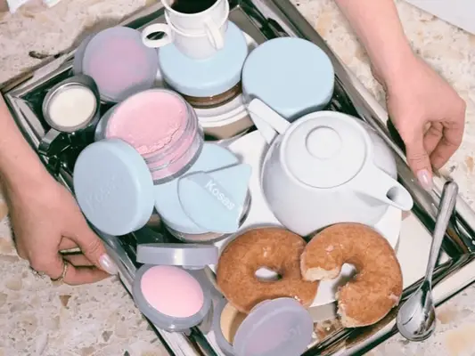 A set of hands placing down a room service tray, which has loose and pressed powders along with a teapot, coffee cups, and donuts