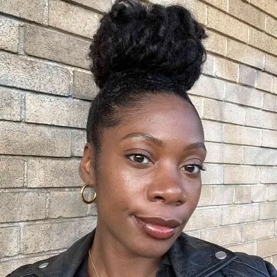 Jasmine Phillips standing against a brick wall, hair pulled back and wearing burgundy lipstick