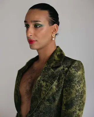 Cyrus Veyssi, a non-binary Iranian person, posing while showing a side profile. They have green eyeshadow on, red lip tint, hair slicked back, and are wearing a velvety green blazer with no shirt on underneath allowing chest hair to show.