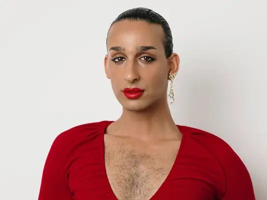 Cyrus Veyssi, a nonbinary Iranian person, wearing a deep-v neckline red dress, red lipstick, eyeliner, and gold dangle earrings. Their hair is slicked back and chest hair is showing through the dress. 