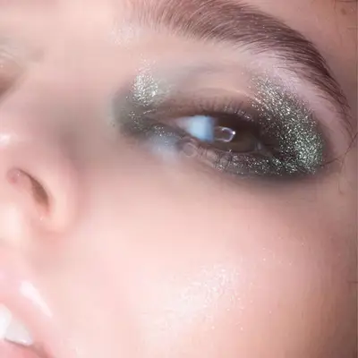 Model wears smoky green glitter eye makeup on inner and outer corners