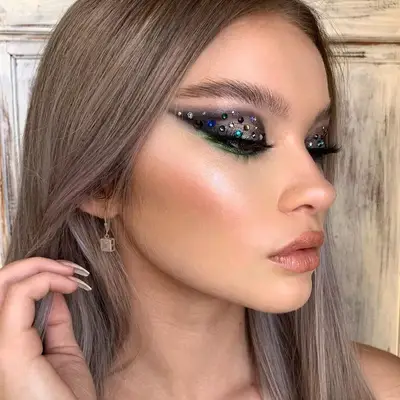 Model with charcoal graphic eyeshadow, emerald green lower liner, and multicolored eyelid gems