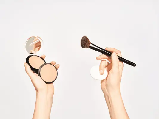 Hands with two powder foundation compacts in one hand and a brush and foundation sponge in the other