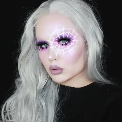 Makeup artist wears purple eyeshadow, blush, and lipstick, long lashes, and white spider web over one eye