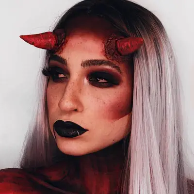 Makeup artist with dark burgundy smoky eye, red contour, black lipstick, and gory devil horns