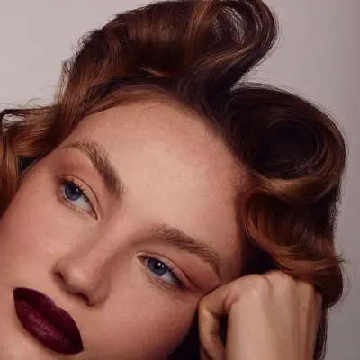 Model with voluminous curled red hair, brushed brows, neutral eyeshadow, and burgundy lipstick