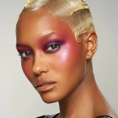 Model wears bright pink blush, purple eyeshadow, and nude glossy lips