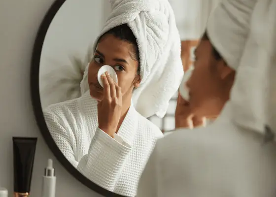 woman using cotton round to remove makeup in mirror