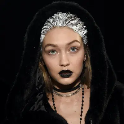 Gigi Hadid wears matte black lipstick, black fur hooded coat, and white paint in hair