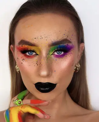 model is wearing rainbow themed makeup on the eyes with paint splatter on the face as an accent. Colors present are red, orange, yellow, violet, blue and green