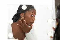 Woman applying HUDA BEAUTY GloWish Multidew Vegan Skin Tint Foundation to her face while looking into mirror