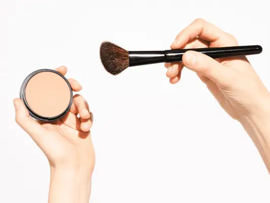 Close up of a set of hands holding a fluffy makeup brush and beige powder compact.