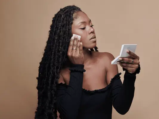 A woman looking at a makeup compact as she applies foundation to her face with a sponge