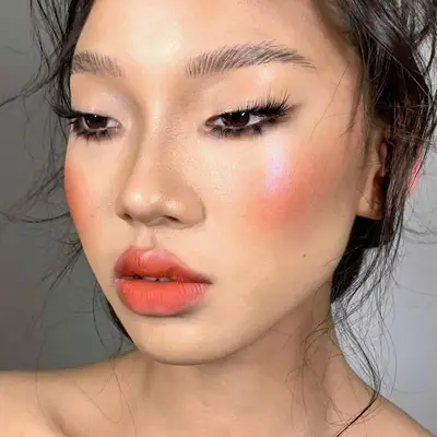 Model wears brushed eyebrows, dramatic lashes and blush, and blended peach lipstick