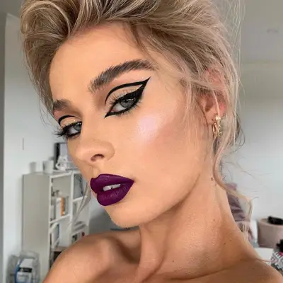 Model wears graphic eyeliner, purple lipstick, and highlighted skin