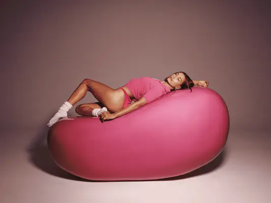 Hailey Bieber wearing a pink polo and pink bikini bottoms laying on a large pink jellybean 