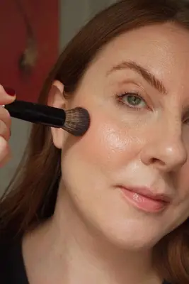 woman applying blush to her cheeks, which are already bronzed