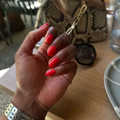 A hand with short red nails with a bag and sunglasses in the background