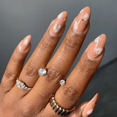 A hand with nude manicured nails and gold stars, ring decorations visible