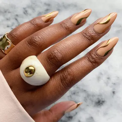 A hand with manicured nude nails featuring a wavy design and wearing gold and white rings, placed on a marble surface