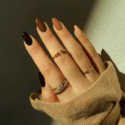 A hand displaying manicured nails in different nude shades, adorned with rings and partially covered by a sweater sleeve