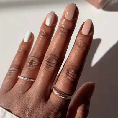 A manicured hand with painted nude nails in different finishes against a light background, adorned with rings on the fingers