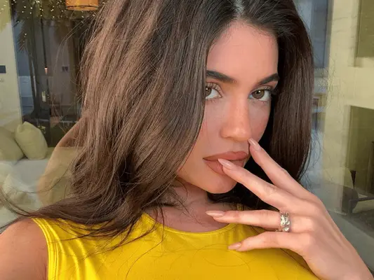 Kylie Jenner in a yellow top holding her almond-shaped nails in a pinky nude color up to her lips