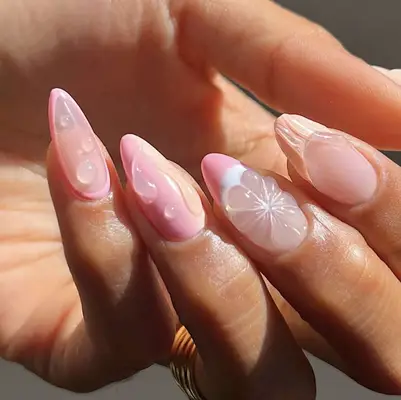 Close-up of a hand with long, artistic fingernails featuring floral and abstract designs on lightly polished surfaces, the nails are of an almond shape and have intricate embellishments