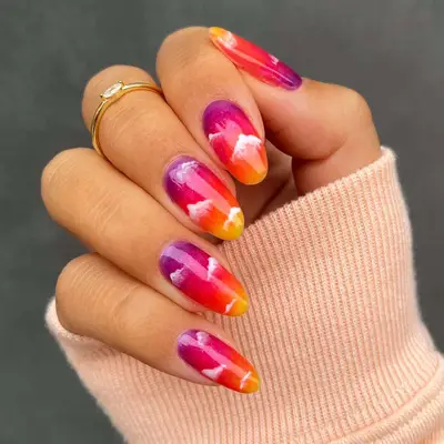 a hand with a purple, red, orange, and yellow sunset and white clouds manicure