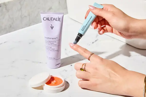Collage of Caudalie and Dior cuticle repair creams on a table beside a person applying Butter London balm to their nail bed