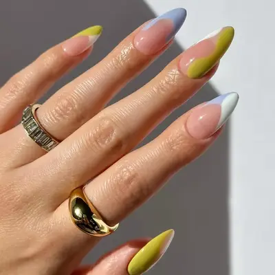 A hand with long, almond-shaped nails featuring a multicolored asymmetric design and two rings on the fingers