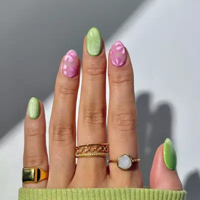 A hand featuring a manicure with green and pink nail designs, accompanied by gold and gemstone rings