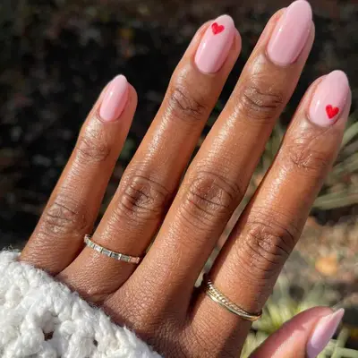 close up of hand showing off fresh manicure, light pink base with small red hearts on two nails