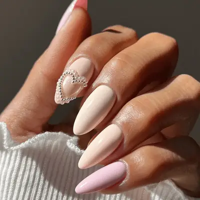 A hand with manicured nails, pale pink polish, and a small rhinestone heart design on one nail
