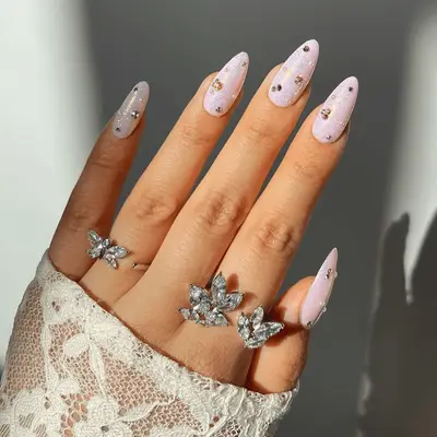 A hand with jewel-adorned pink nails and decorative rings, wearing lace