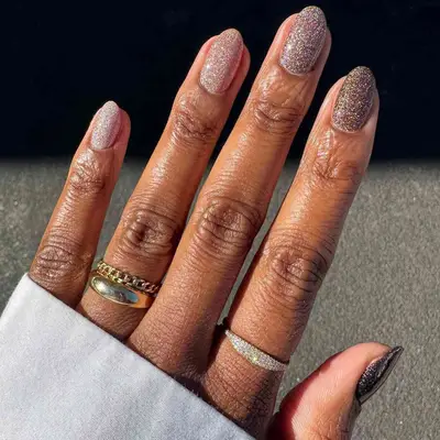 A hand with neatly manicured neutral glittery nails, wearing rings on the fingers, against a textured surface background