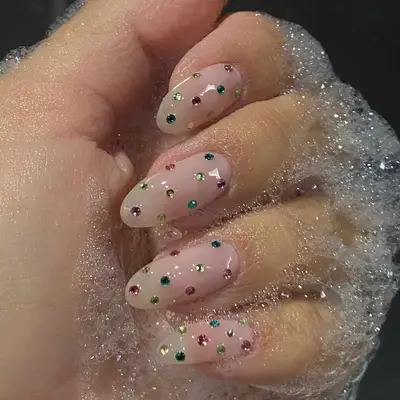 Hand with glossy nail art featuring small multicolored rhinestones, surrounded by soap suds