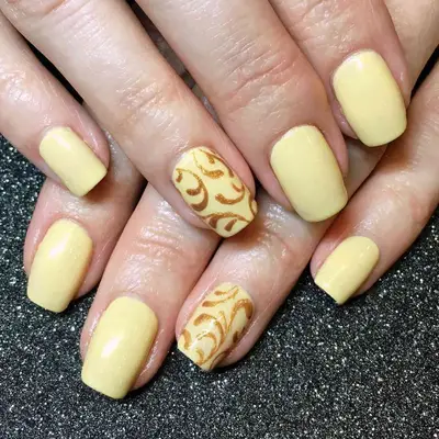 Butter yellow nails with gold floral detailing on the ring finger.
