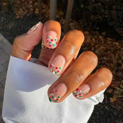 A neutral manicure with white, red, green, and silver polka dots at the tips
