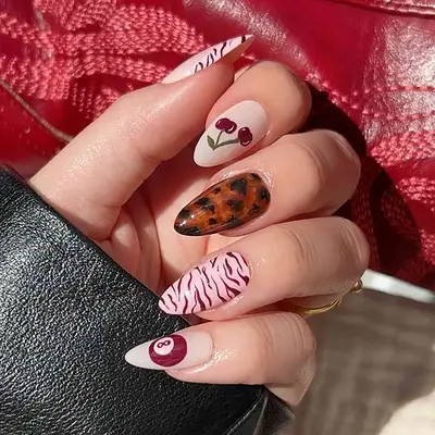 A close-up of a hand with nail art featuring designs such as cherries, a tortoiseshell pattern, zebra stripes, and an 8-ball, holding a leather strap
