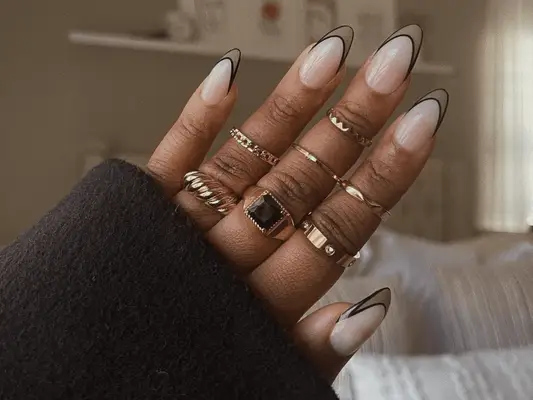 A hand with rings on the three middle fingers and press-on nails that have a negative space French tip with black lines